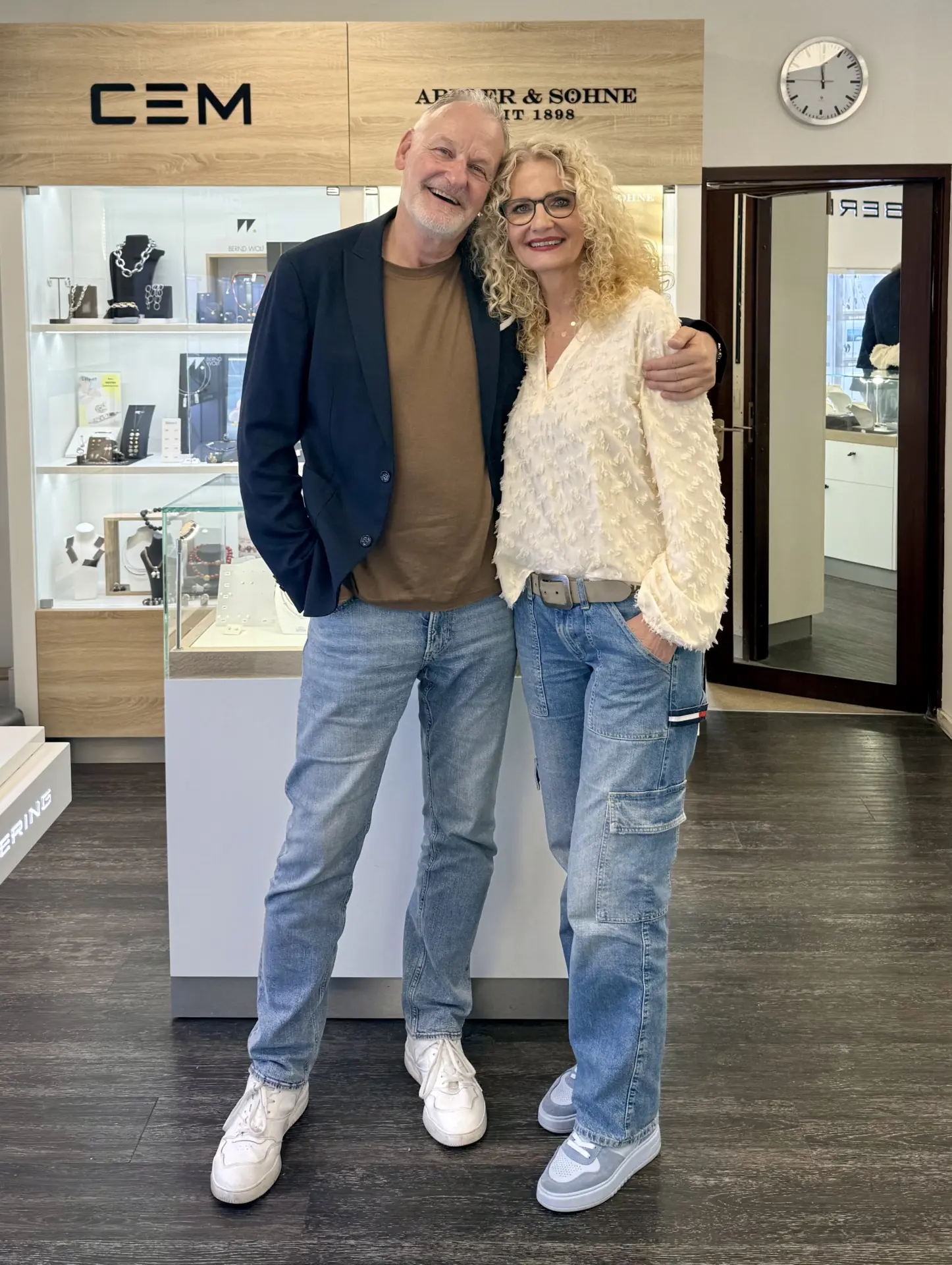 Gudrun Lehmkühler und Matthias Lehmkühler im Juwelier Lehmkühler Store Hamm, blonde Haare, Jeans-Outfit, Brille, freundliches Lächeln vor Schmuckauslage